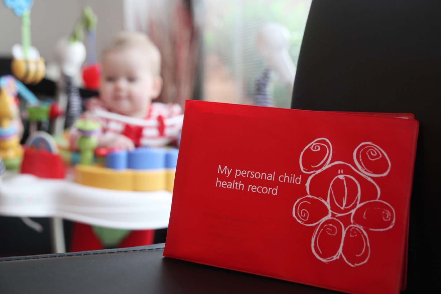 A red personal child health record booklet in front of a baby